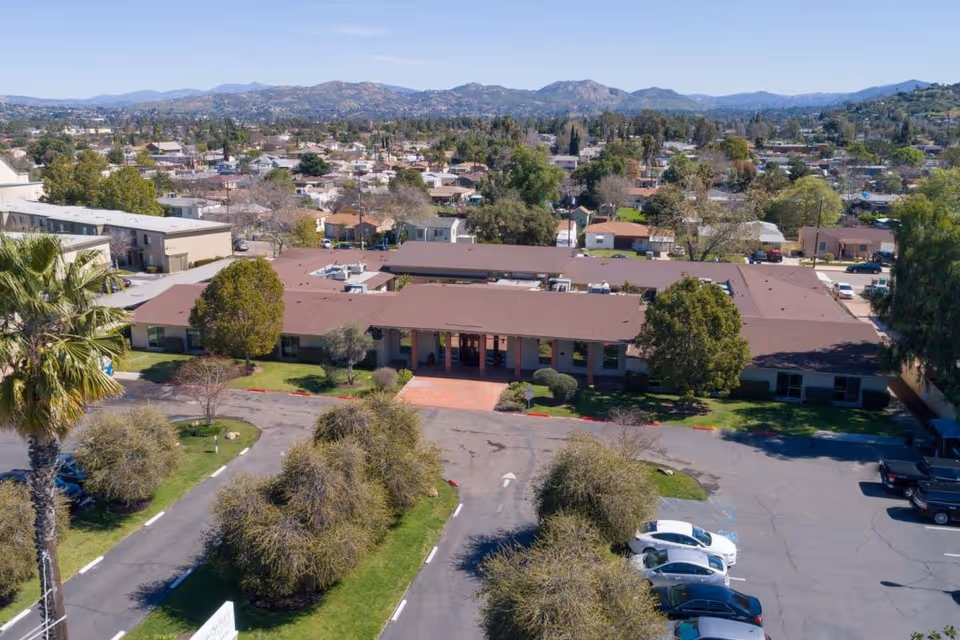 Aerial view of Magnolia Post Acute Care facility showing a single-story building with a brown roof surrounded by trees and a parking lot with several cars. The background includes a suburban neighborhood and distant mountains under a clear blue sky.