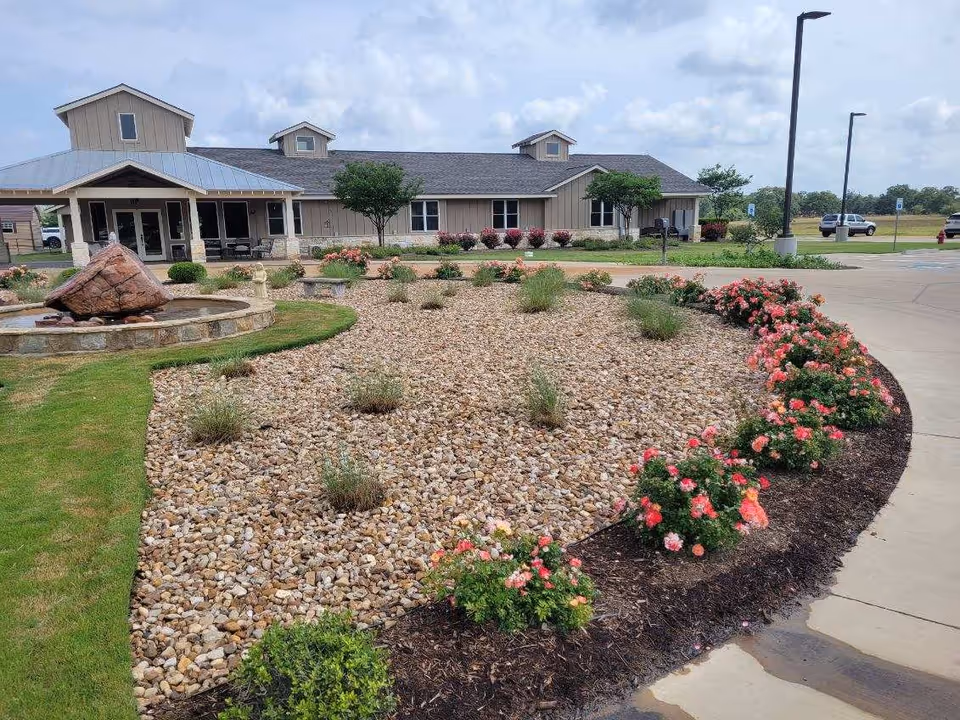 Exterior view of Bec's Blessing facility showing a landscaped garden with a rock fountain, flowering bushes, and small trees in front of a single-story building with a metal roof. The sky is partly cloudy and there is a paved driveway and parking area visible.