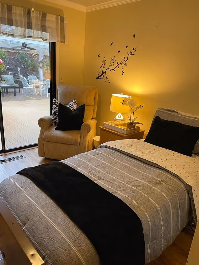 A cozy bedroom with a single bed covered in gray and black bedding, a beige armchair with two decorative pillows, a wooden nightstand with a lamp and a small plant, and a wall decal of a tree branch with birds. A sliding glass door opens to an outdoor patio with white metal chairs and a table.