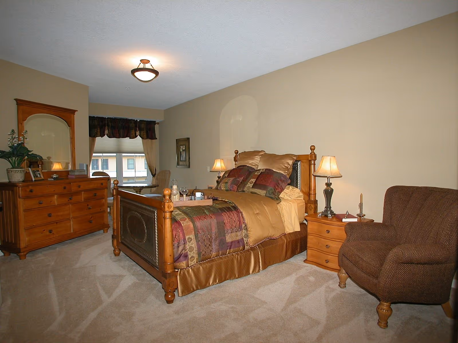 A cozy bedroom with a wooden bed frame, a colorful patterned bedspread, and multiple pillows. There is a wooden nightstand with a lamp and a candle holder, a brown upholstered armchair, and a wooden dresser with a mirror and decorative items. A window with curtains is visible in the background.