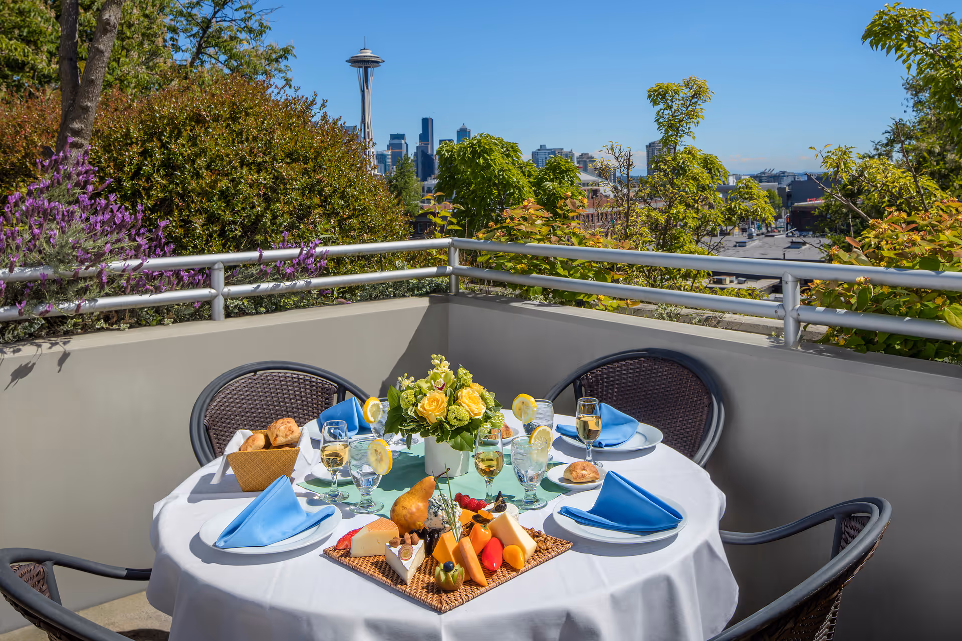 Outdoor dining table set on a balcony with a white tablecloth, blue napkins, glasses with lemon slices, a basket of bread rolls, a cheese and fruit platter, and a vase of yellow roses. The balcony overlooks greenery and a city skyline with the Space Needle visible in the background under a clear blue sky.