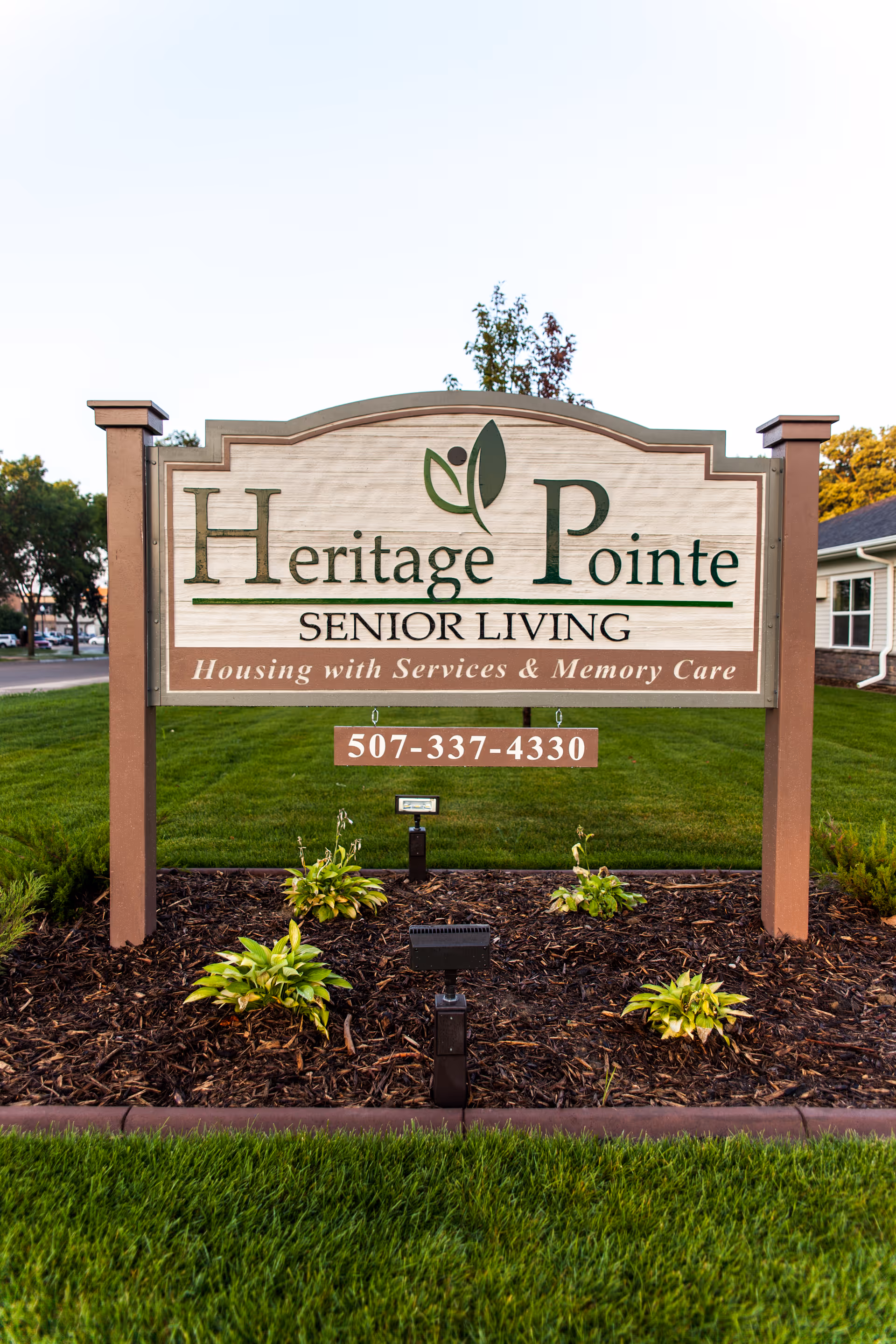 A wooden sign for Heritage Pointe Senior Living displayed outdoors on a landscaped area with green grass and small plants. The sign includes the text 'Housing with Services & Memory Care' and a contact phone number 507-337-4330.