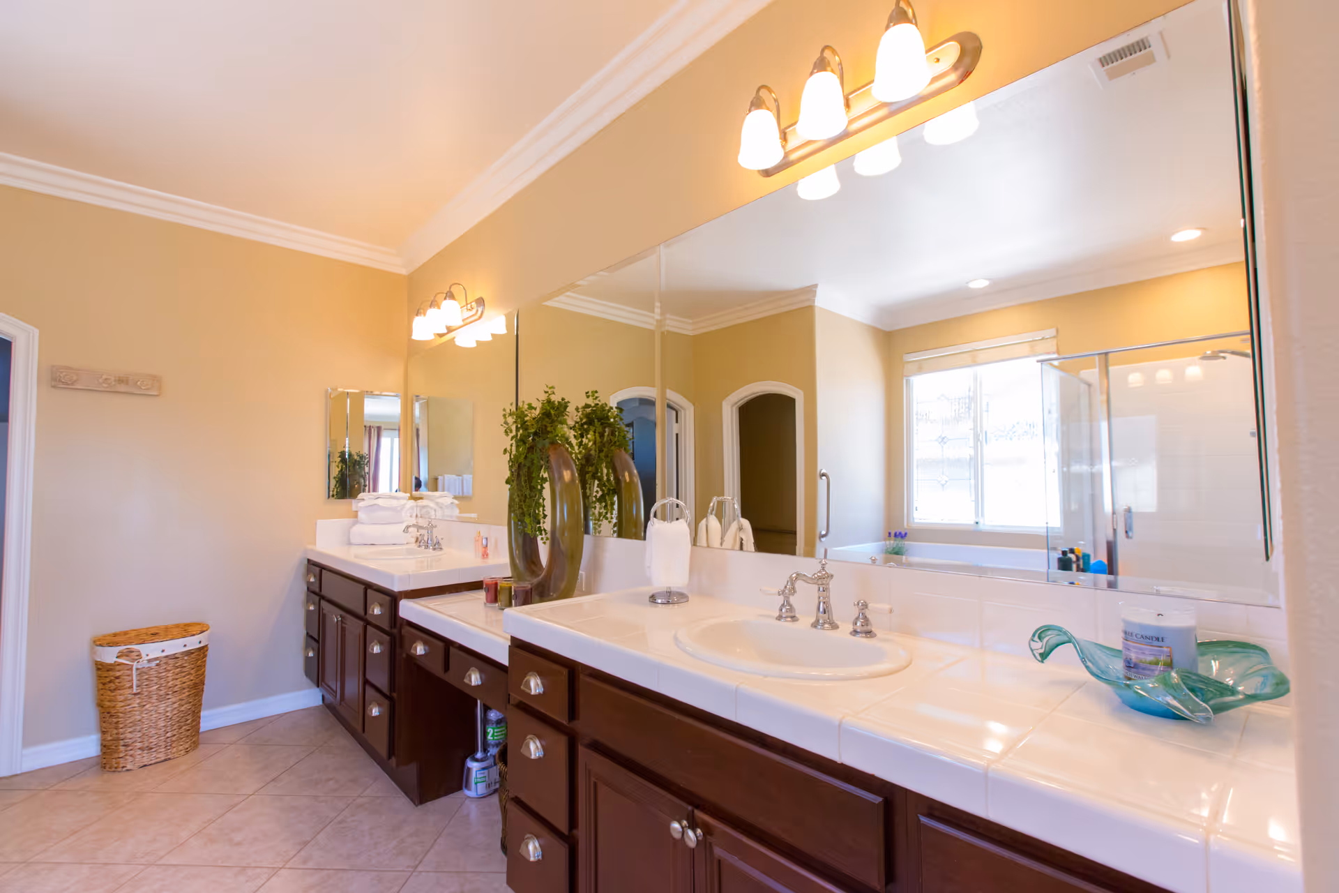 A spacious bathroom with beige walls and tiled floor featuring a long countertop with two sinks, dark wooden cabinets underneath, a large mirror above, and decorative items including a plant and a candle. There is a window with blinds and a glass-enclosed shower visible in the background.