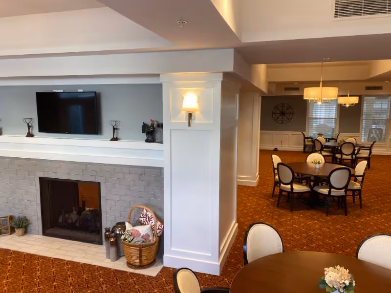 A cozy common area in a senior living facility featuring a fireplace with a mounted TV above it, decorative items on the mantel, and a basket with blankets nearby. The room has multiple round tables with chairs arranged for socializing or dining, warm lighting, and carpeted floors.