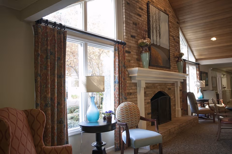 A cozy living room area with a brick fireplace, a white mantel decorated with vases and a framed painting above it. There are patterned curtains on large windows letting in natural light. The room has a mix of upholstered chairs and a small round table with a blue glass lamp and a small potted plant.