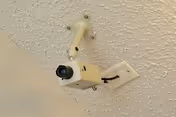 A ceiling-mounted security camera and bracket attached to a textured ceiling and wall outlet.