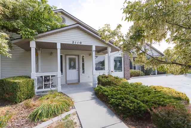 Front entrance of a single-story senior living building with a covered porch, white entry door, and surrounding shrubs and trees.