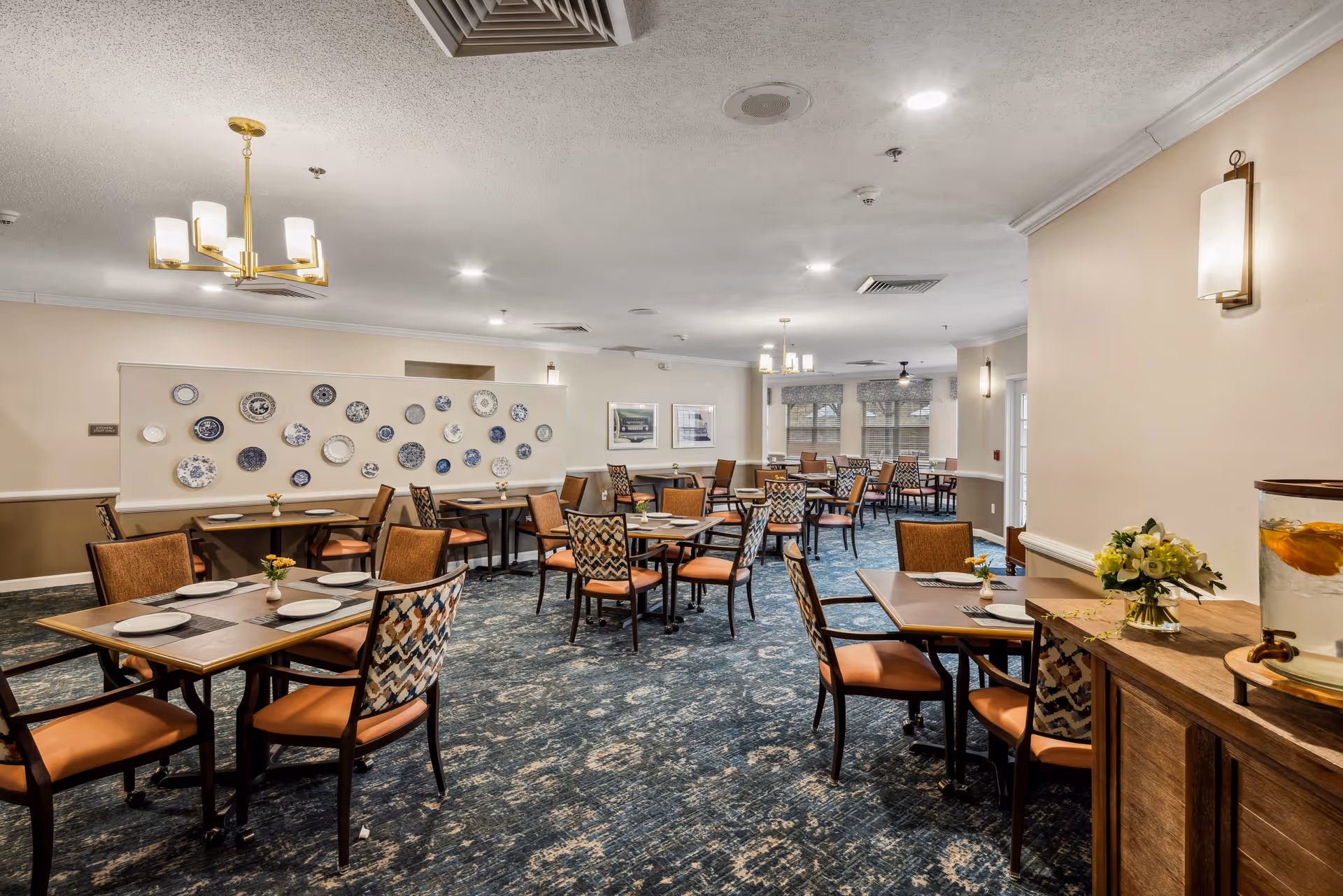 A spacious dining room with multiple tables and chairs arranged neatly. The tables are set with plates and small flower vases. The room features a blue patterned carpet, beige walls, decorative plates on a partition wall, and modern light fixtures hanging from the ceiling. A wooden sideboard with a glass container of infused water and a flower arrangement is visible on the right side.