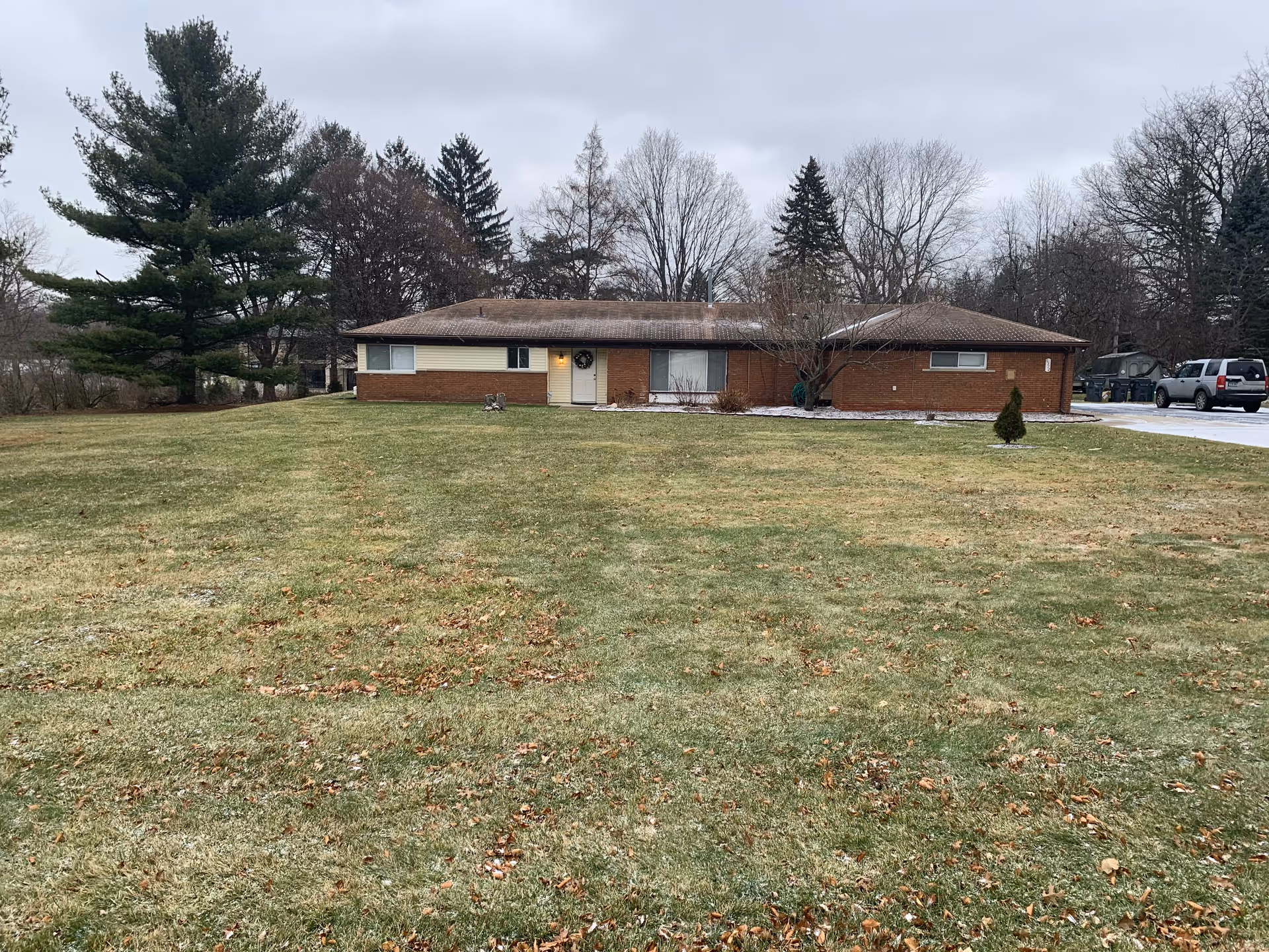 Single-story brick building with a large grassy lawn in front, surrounded by trees under a cloudy sky. There are a few vehicles parked on the right side near the building.