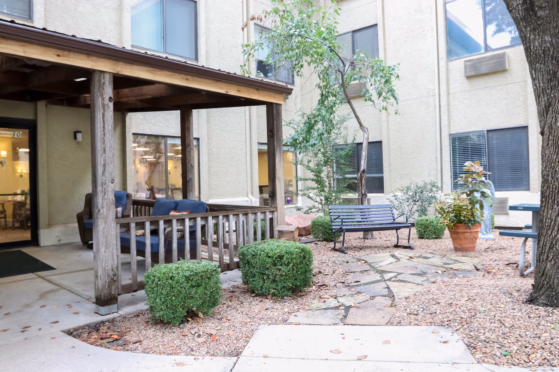 Outdoor courtyard area at Sedgwick Plaza featuring a covered wooden porch with cushioned seating, a stone pathway, trimmed bushes, a metal bench, potted plants, and surrounding building windows.