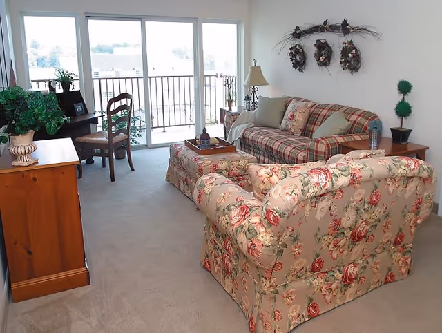 A cozy living room with floral and plaid upholstered furniture, including a floral armchair and a plaid sofa. There is a wooden side table with a lamp and a small plant, a wooden desk with a chair, and a sliding glass door leading to a balcony with a view outside. The walls are decorated with three wreaths and a branch above the sofa.