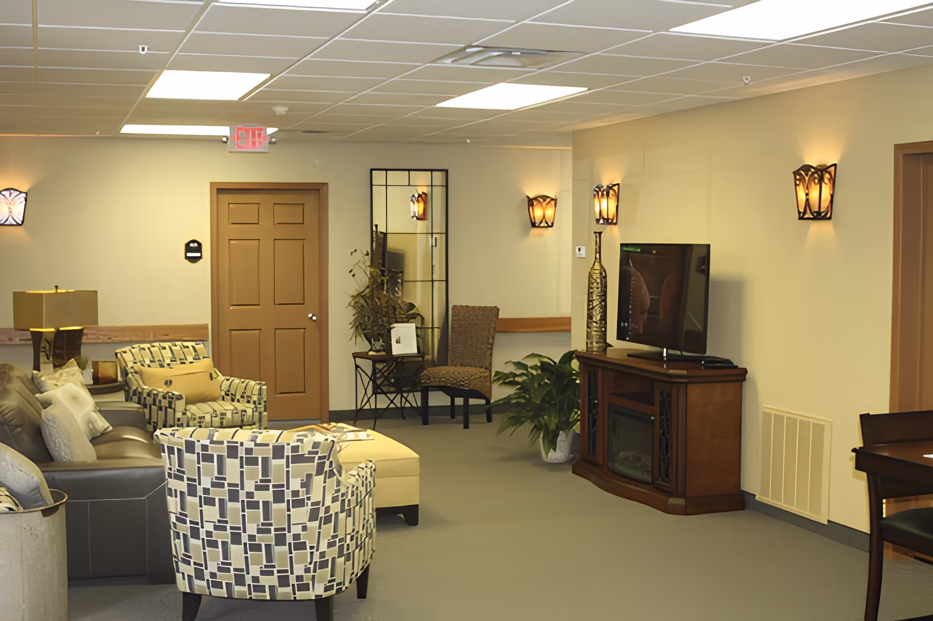 A cozy living room area in a retirement center with patterned armchairs, a leather sofa, a wooden TV stand with a flat-screen television, decorative wall sconces, a large mirror, and indoor plants. The room has a beige carpet and neutral-colored walls with wooden doors.
