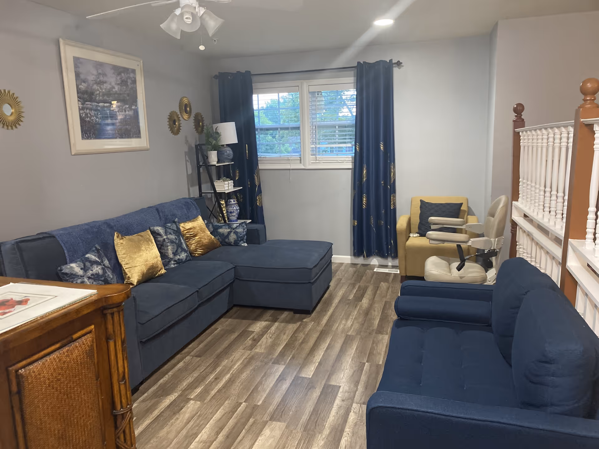 A cozy living room with a blue sectional sofa adorned with gold and patterned pillows, a blue loveseat, a yellow armchair, and a beige mobility chair. The room has wood flooring, a window with blue curtains, a ceiling fan with lights, and decorative wall art including a framed painting and gold sunburst mirrors.