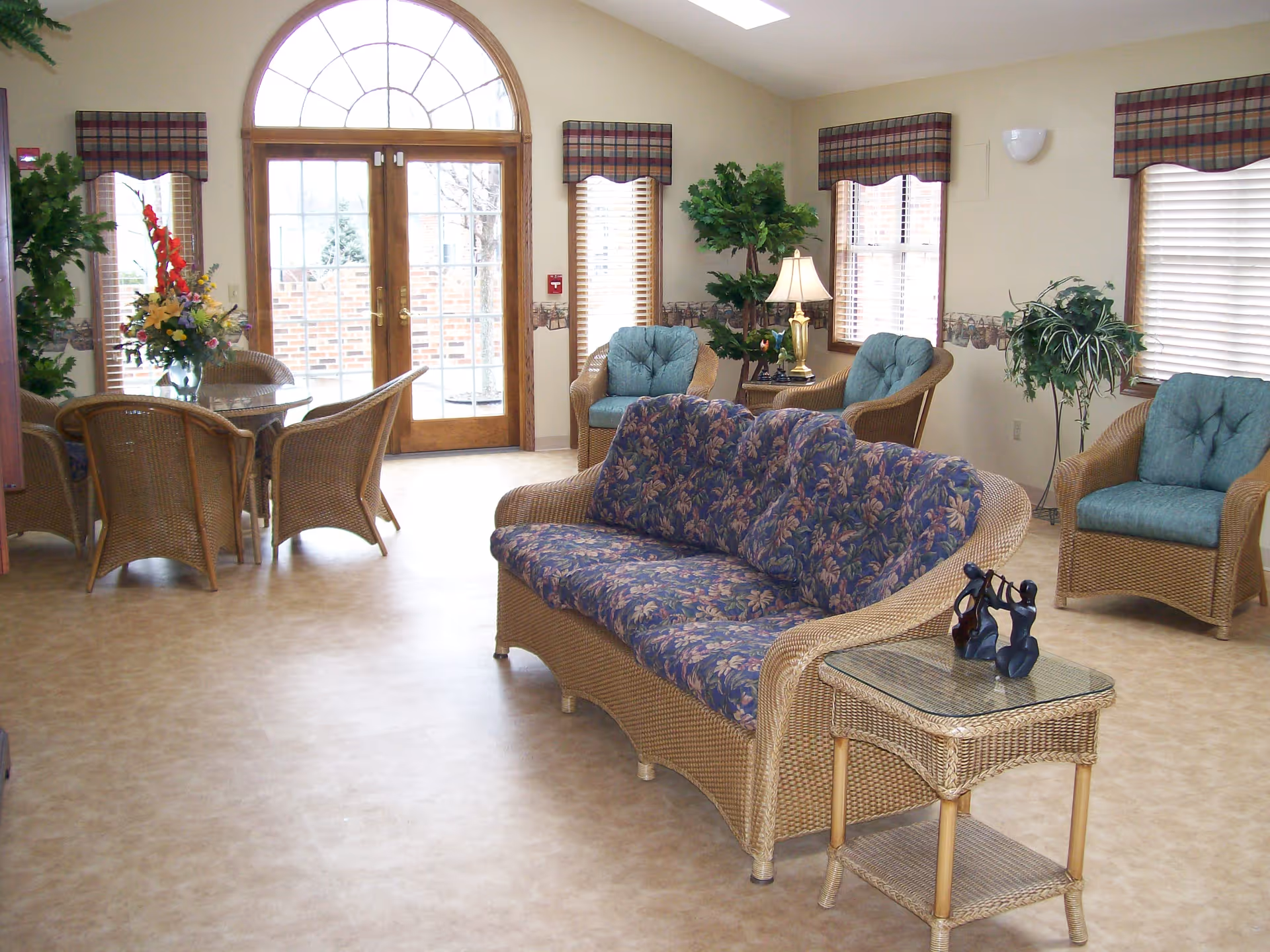 Bright sitting area with wicker sofas and chairs arranged around a glass-top side table and a round dining table near arched double doors.