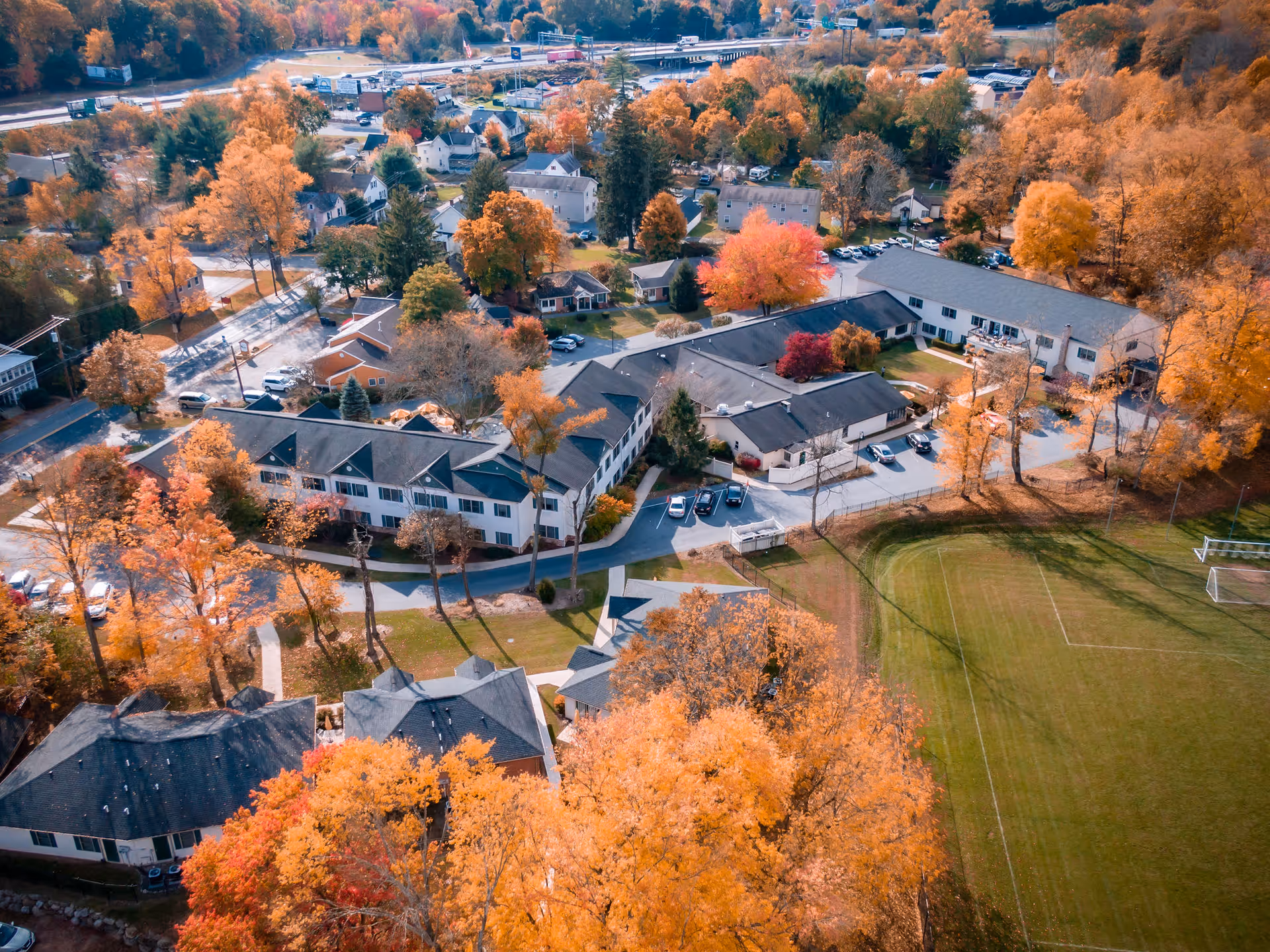 Aerial view of Grace Park Senior Living facility surrounded by autumn trees with vibrant orange, yellow, and red foliage. The facility consists of multiple connected buildings with parking areas and nearby residential houses. A green sports field with soccer goals is visible adjacent to the facility.