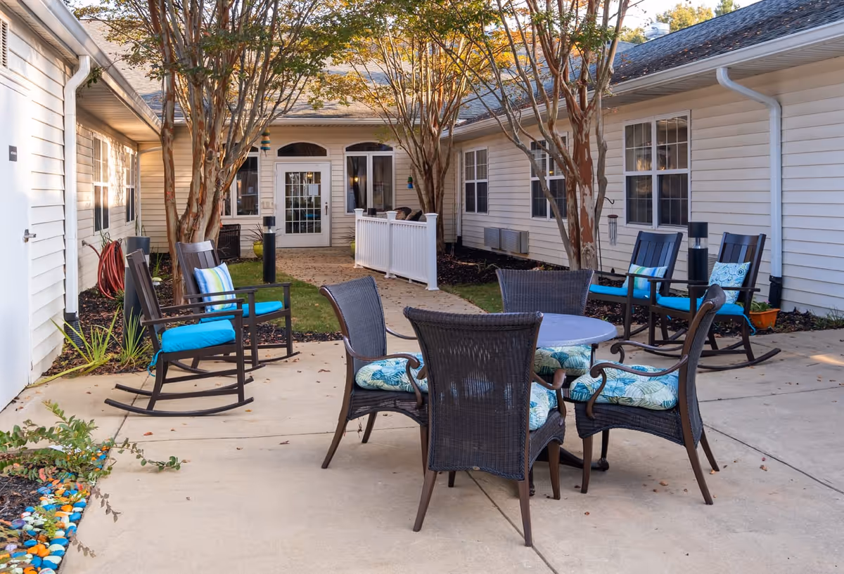 Outdoor patio area at The Pines at Greenville Assisted Living & Memory Care with several black wicker chairs and a round table with cushions, surrounded by white buildings and trees with autumn leaves.