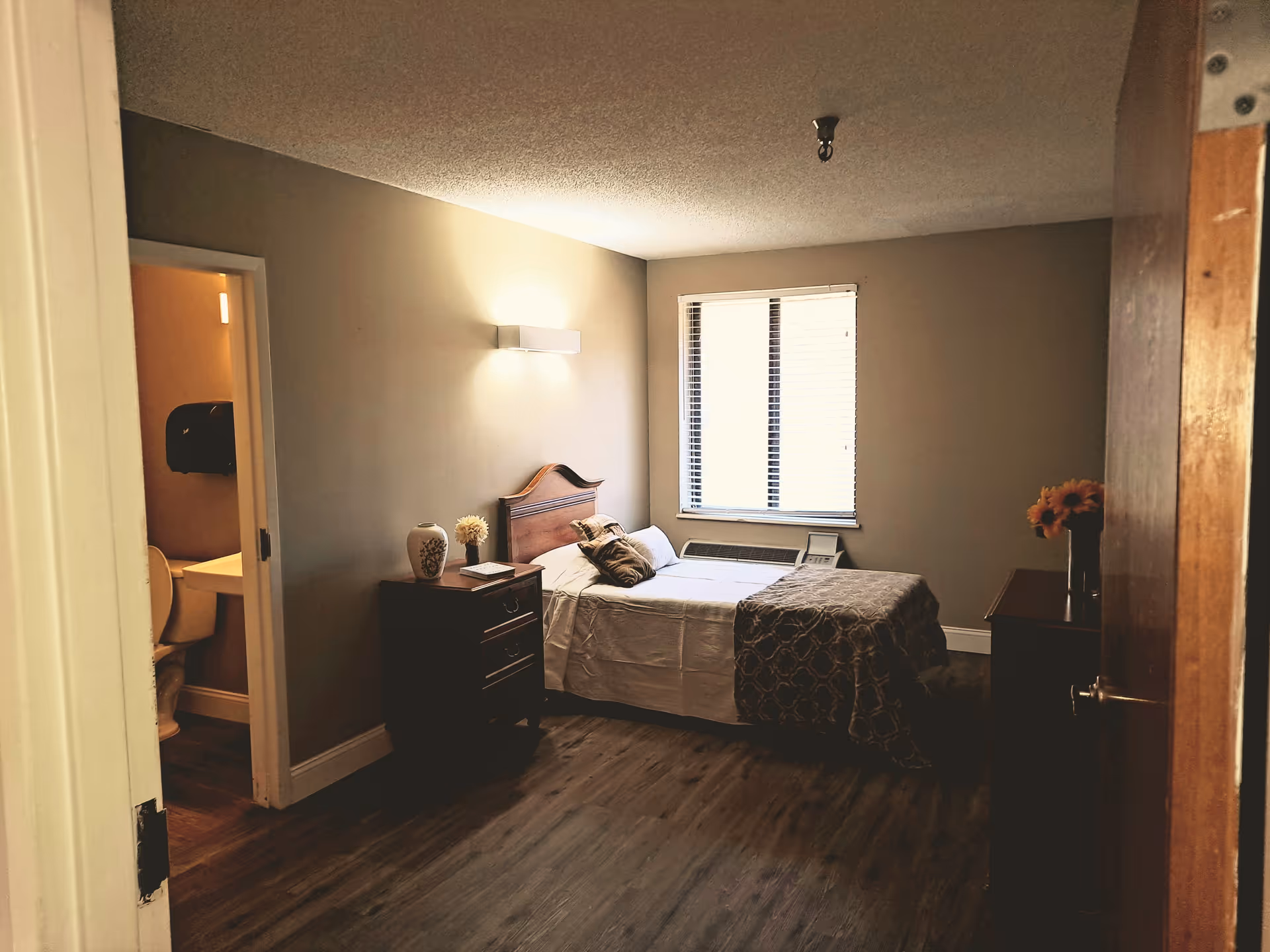 Furnished bedroom with a single bed, nightstand and dresser, a window, and an open doorway to a bathroom.