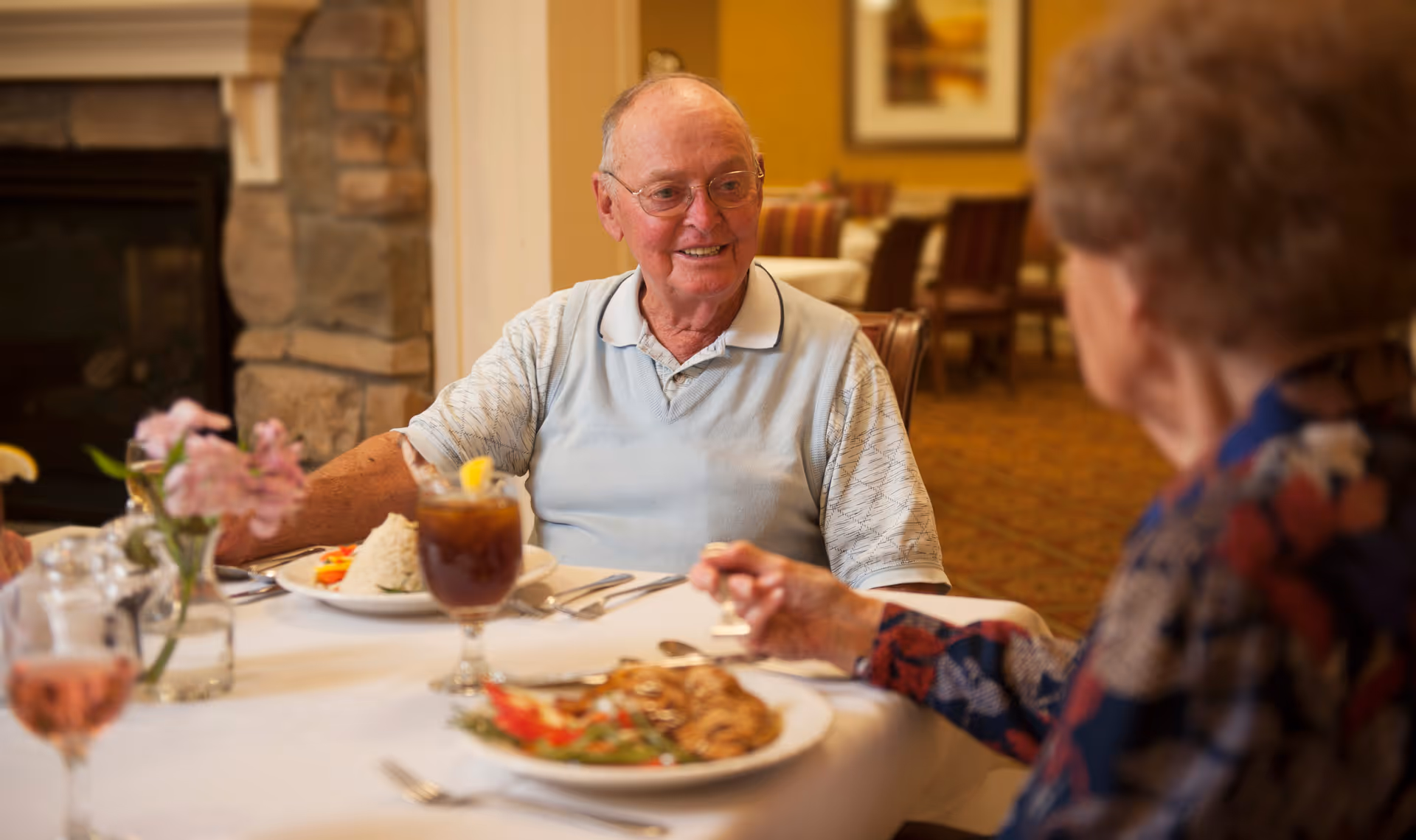 An elderly man and woman are seated at a dining table in a warmly lit room. The man is smiling and wearing glasses and a light blue polo shirt. On the table are plates of food, glasses of iced tea and wine, and a small vase with pink flowers. The background shows more dining tables and chairs in a cozy setting.