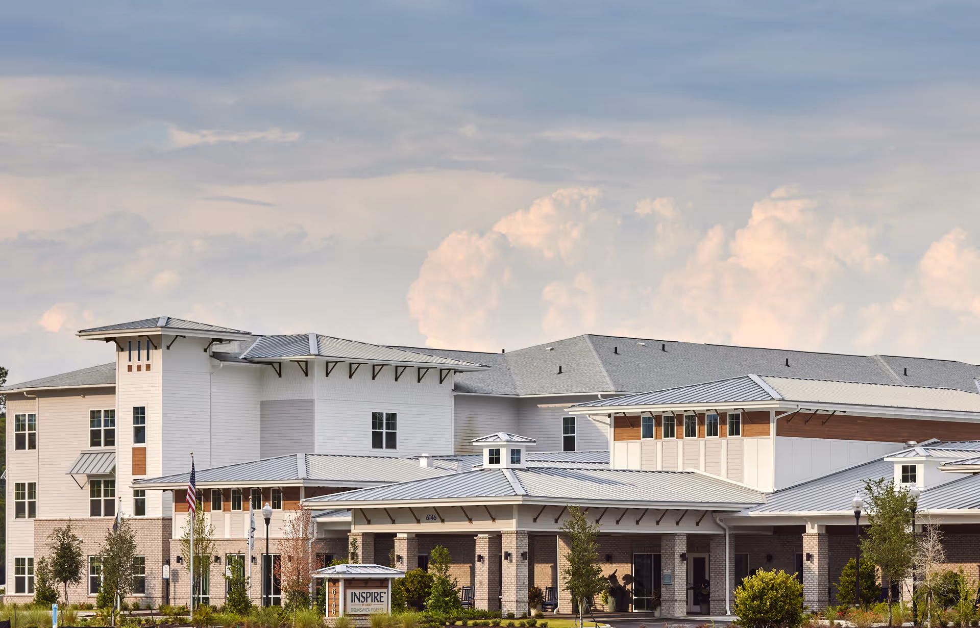Front exterior of the Inspire Brunswick Forest senior living building with multiple gabled roofs and a landscaped entrance under a cloudy sky.
