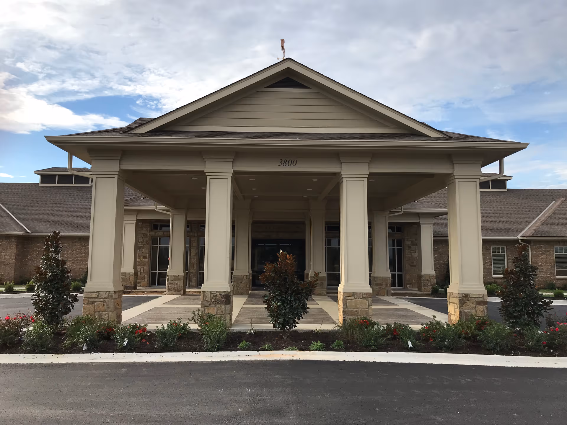 Front exterior view of a senior living facility building with a covered entrance supported by columns, landscaped with bushes and flowers, under a partly cloudy sky.