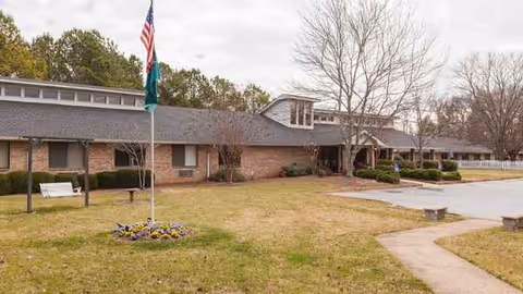 Single-story assisted living facility with flagpole, brick facade, and manicured lawn.