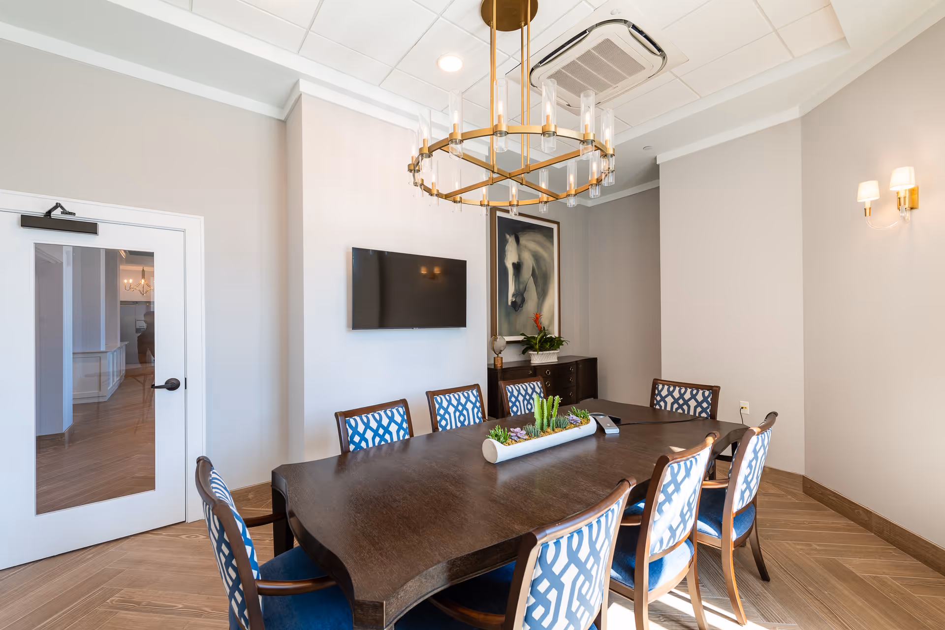 A well-lit dining room with a dark wooden table surrounded by eight chairs with blue and white patterned upholstery. A modern chandelier hangs above the table. On the wall, there is a flat-screen TV and a framed painting of a white horse. A small cabinet with a plant and decorative items is placed against the wall. The room has light-colored walls and wooden flooring.