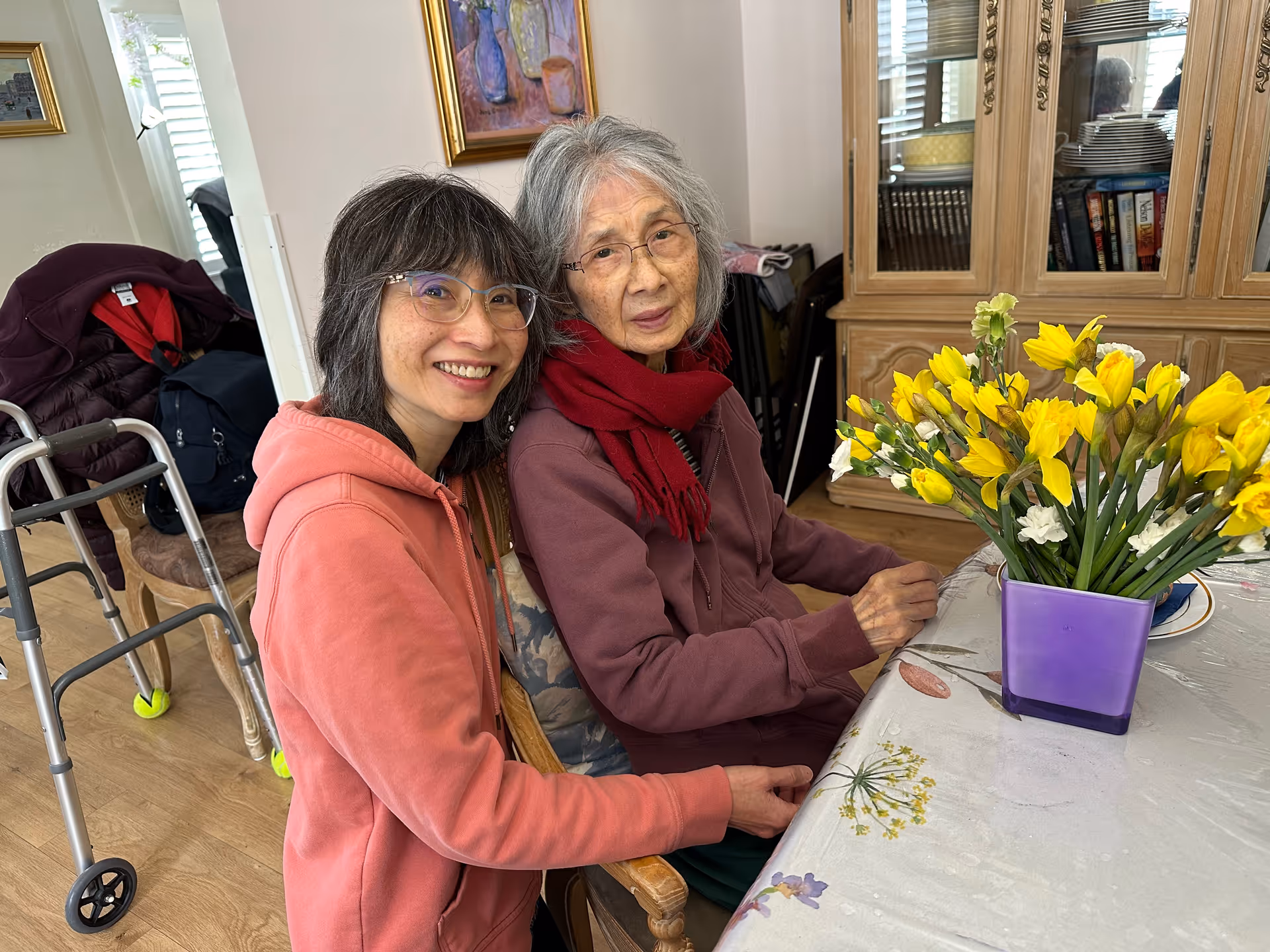 Two women sitting at a table indoors, one elderly woman wearing glasses and a red scarf, and a younger woman with glasses and a pink hoodie smiling beside her. On the table is a purple vase filled with yellow flowers. In the background, there is a wooden cabinet with glass doors and some framed artwork on the wall.