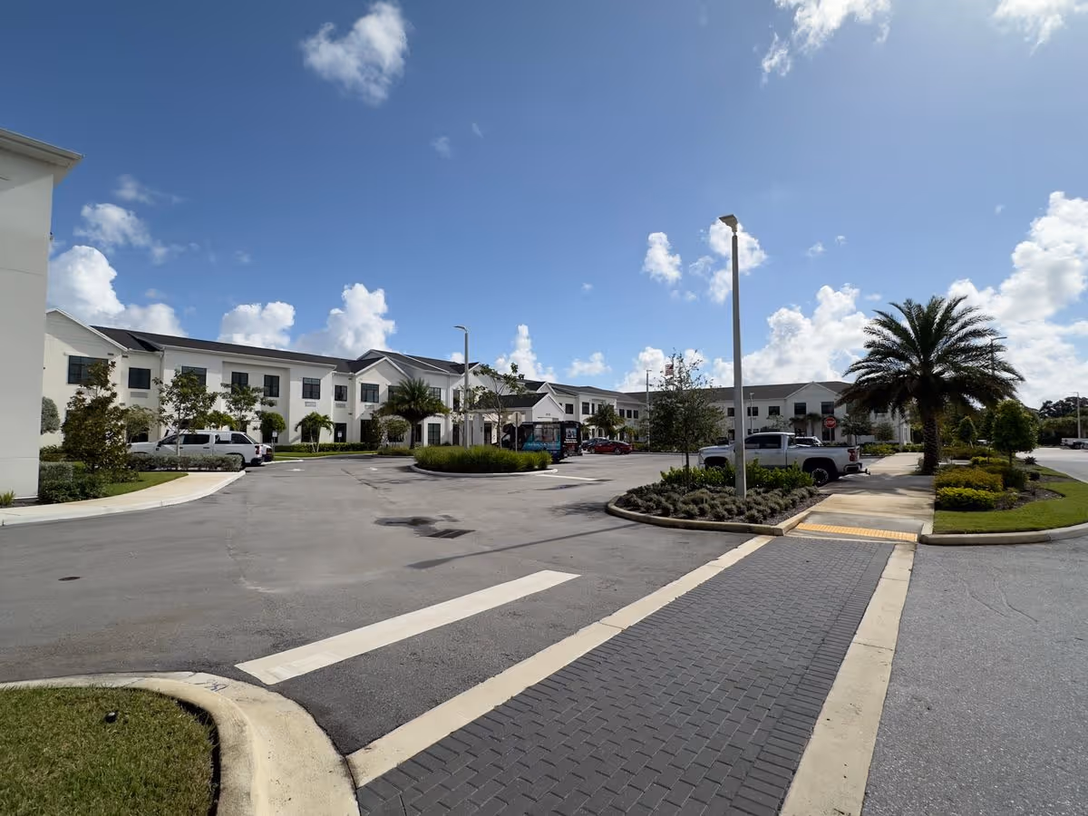 Front view of a two-story white building with a circular driveway, palm trees, and parked cars under a blue sky.
