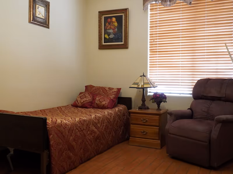A cozy bedroom with a single bed covered in a red patterned bedspread and matching pillows. Next to the bed is a wooden nightstand with two drawers, a decorative lamp, and a small flower arrangement. A large brown recliner chair is positioned near a window with closed wooden blinds. Two framed pictures hang on the beige walls.