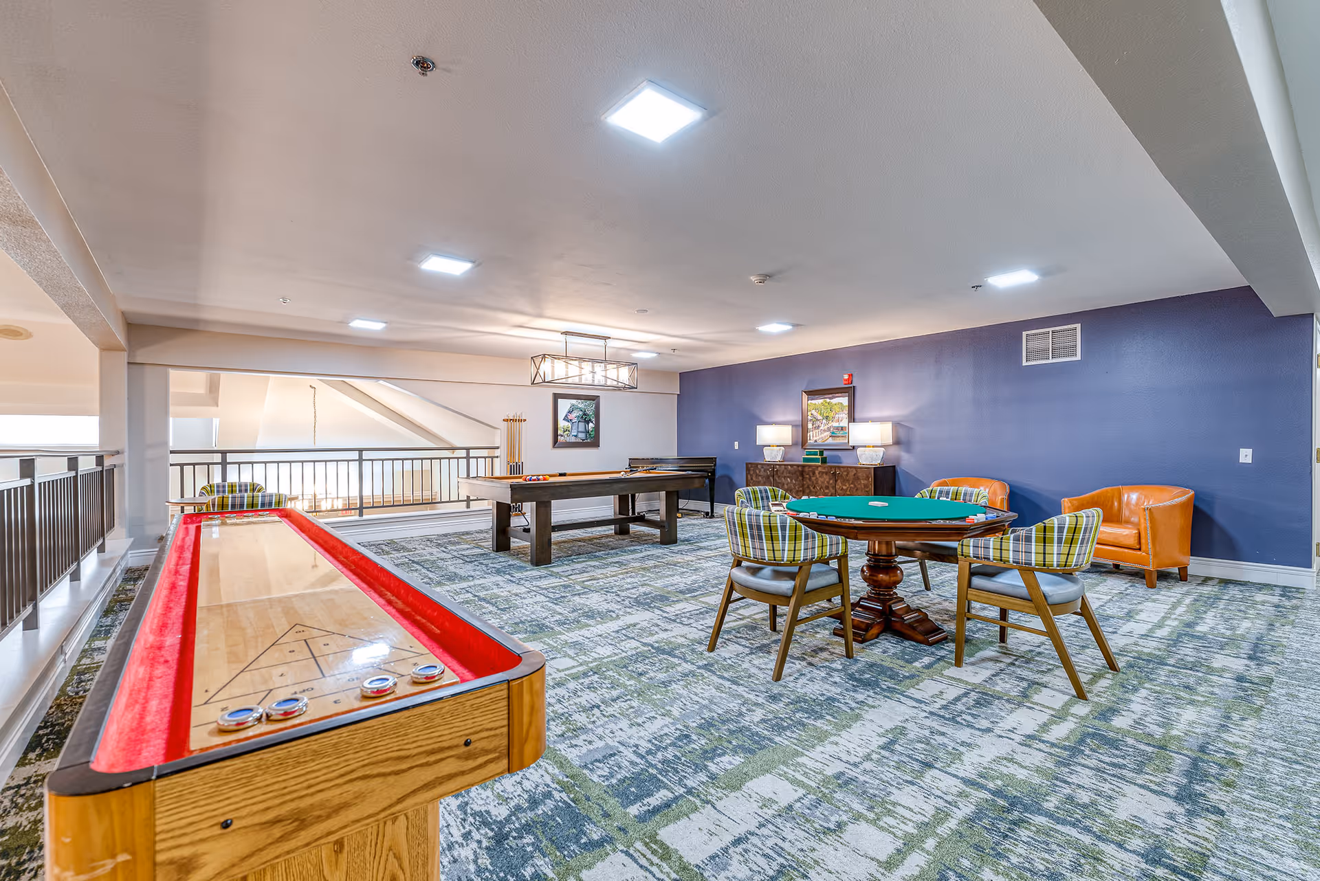 A bright senior living game room with a shuffleboard table, pool table, poker table with chairs, and seating against a blue accent wall.