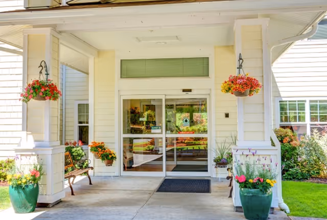 Entrance to a senior living facility with a covered porch area featuring hanging flower baskets, potted plants, a bench, and glass automatic sliding doors leading inside.