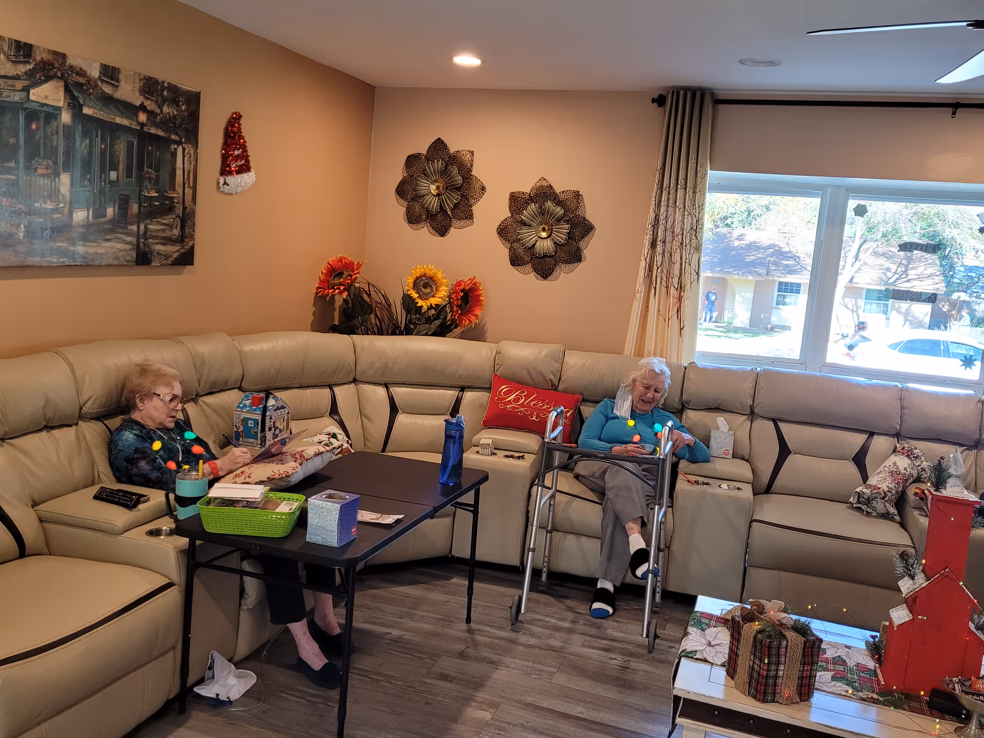 Two elderly women sit on a beige sectional sofa in a cozy, decorated living room with a coffee table and holiday decorations.