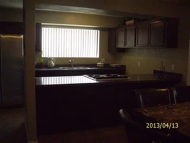 Dimly lit kitchen with a central island and cooktop, sink under a window with vertical blinds and dark cabinets.