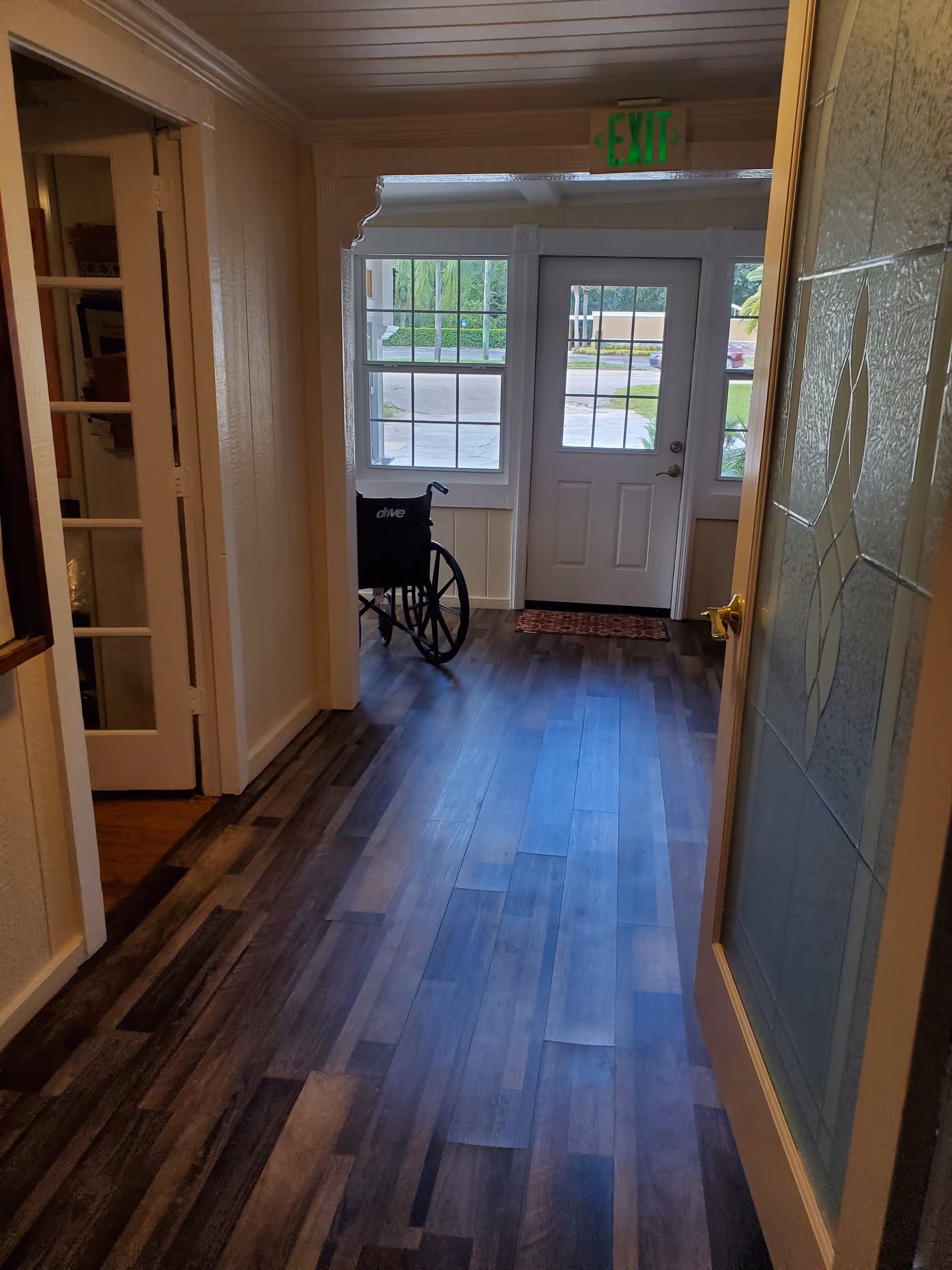Interior hallway with wood flooring leading to a door with glass panels and windows on either side. A wheelchair is positioned near the windows. There is an exit sign above the door and a decorative glass panel on the right side of the hallway.