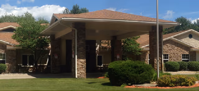 Exterior view of a senior living facility building with stone and siding walls, a covered entrance supported by stone pillars, a well-maintained lawn, bushes, and trees under a partly cloudy blue sky.
