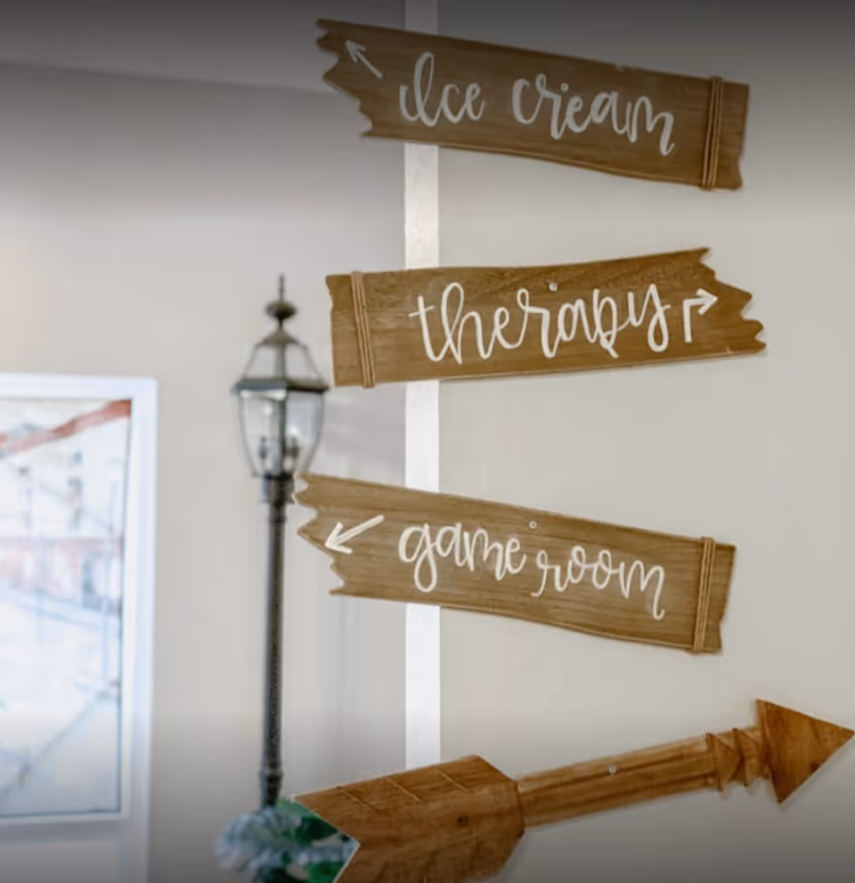 Wooden directional signs mounted on a wall pointing to ice cream, therapy, and game room, with a decorative street lamp and a window in the background.