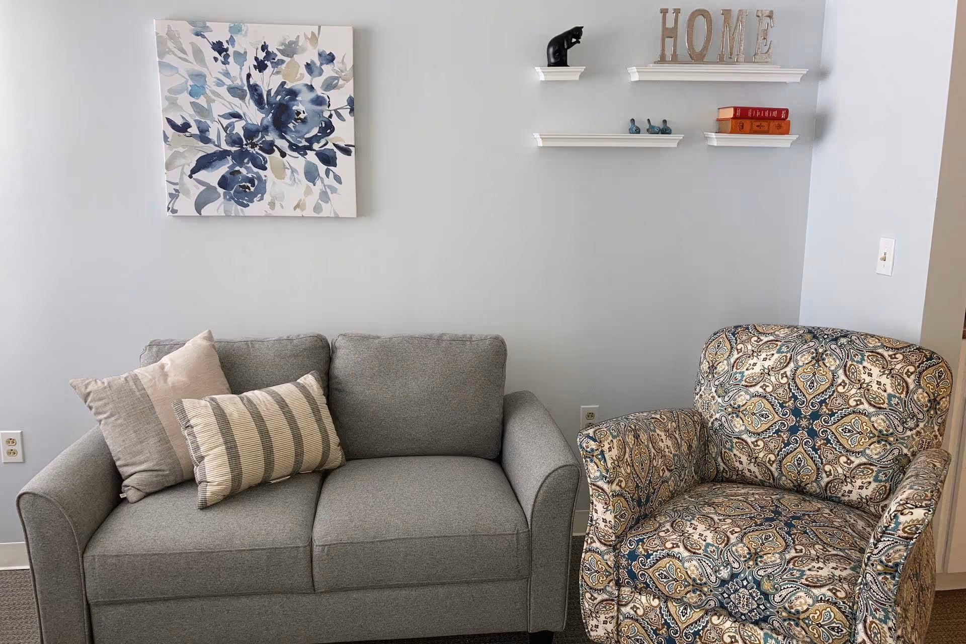 A cozy living area with a gray loveseat adorned with three pillows and a patterned armchair. Above the loveseat, there is a floral painting with blue and gray tones. On the adjacent wall, three white floating shelves hold decorative items including a black cat figurine, small blue bird figurines, two orange books, and wooden letters spelling 'HOME'.