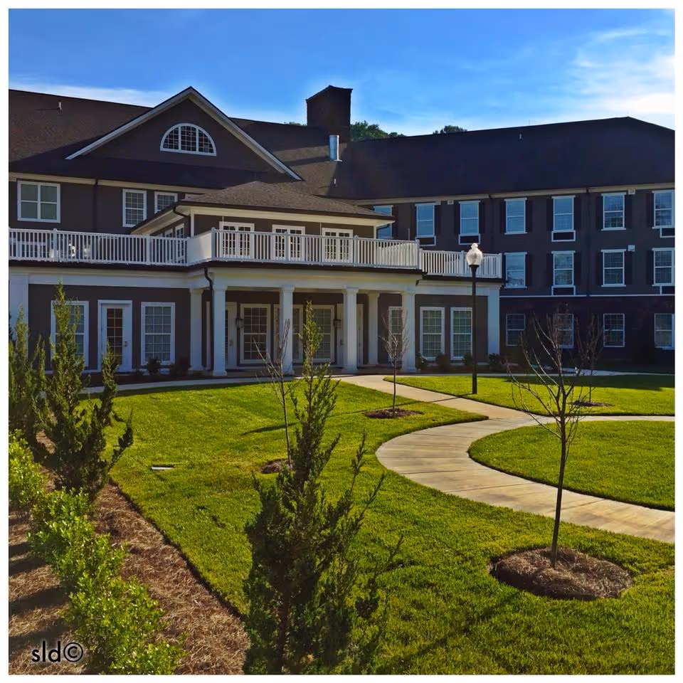 Exterior view of a senior living facility building with multiple windows, a balcony, and a covered porch supported by columns. The foreground features a well-maintained lawn with a curved concrete walkway, young trees, and shrubs under a clear blue sky.