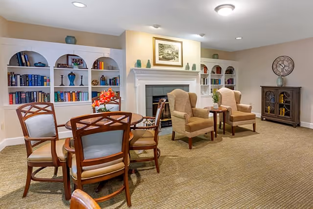 A cozy common area with a round wooden table surrounded by four chairs in the foreground. In the background, there are two beige armchairs with a small wooden side table between them, placed in front of a white fireplace with decorative items on the mantel. Built-in white bookshelves filled with books and decorative objects flank the fireplace. A wooden cabinet with a clock on top is positioned against the wall on the right.