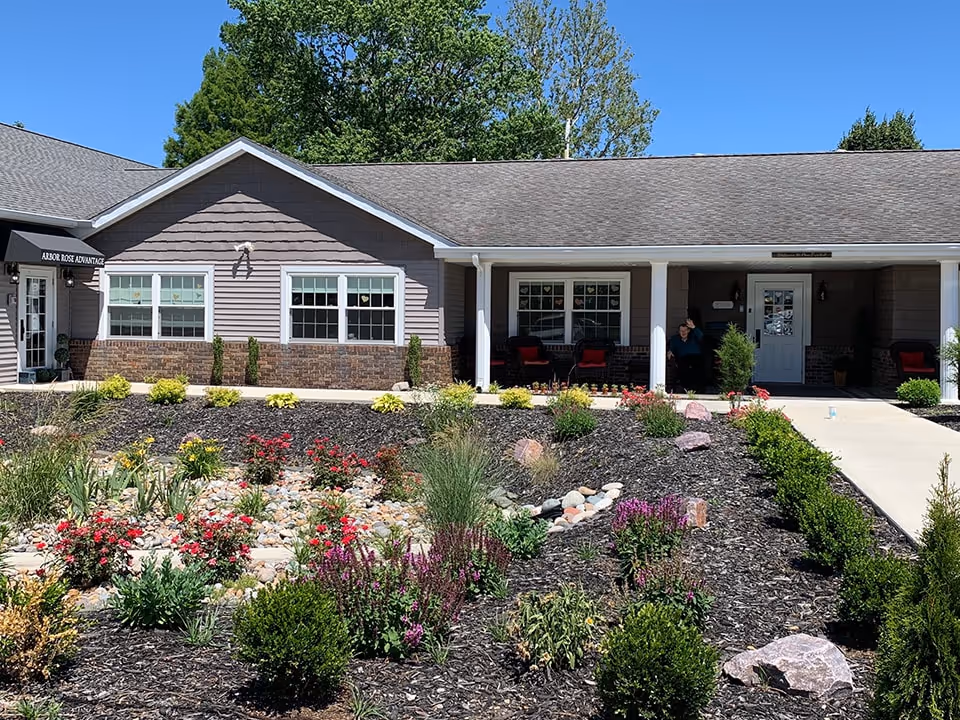 Exterior view of Arbor Rose of Monticello facility showing a single-story building with a covered porch, several windows, and a landscaped garden with various plants and flowers in the foreground under a clear blue sky.