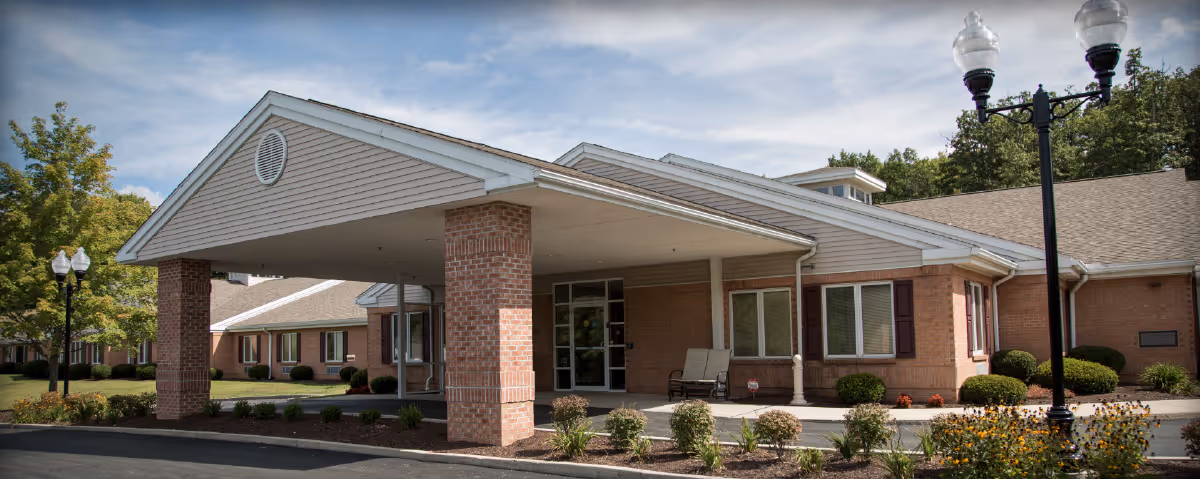 Front entrance of a brick rehabilitation and care center with a covered porte-cochere, landscaped beds, and lampposts.