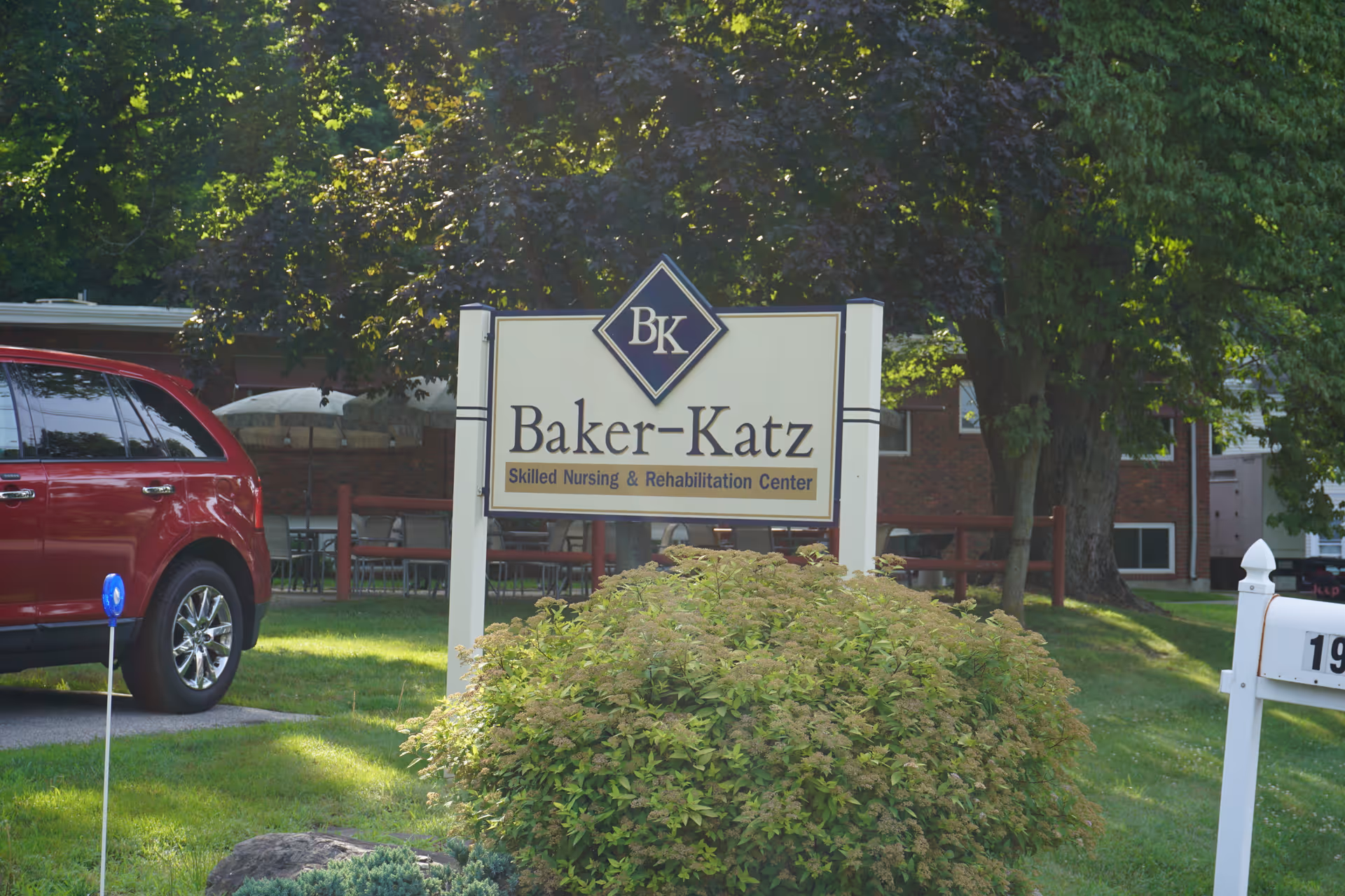 Entrance sign that reads 'Baker-Katz Skilled Nursing & Rehabilitation Center' standing on a lawn with a building and trees behind it.