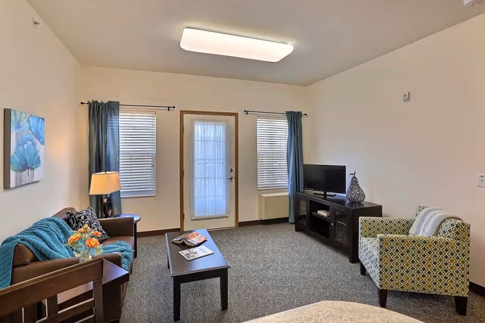 A bright furnished living room with a sofa, patterned armchair, coffee table, TV on a console, and a door flanked by two windows.