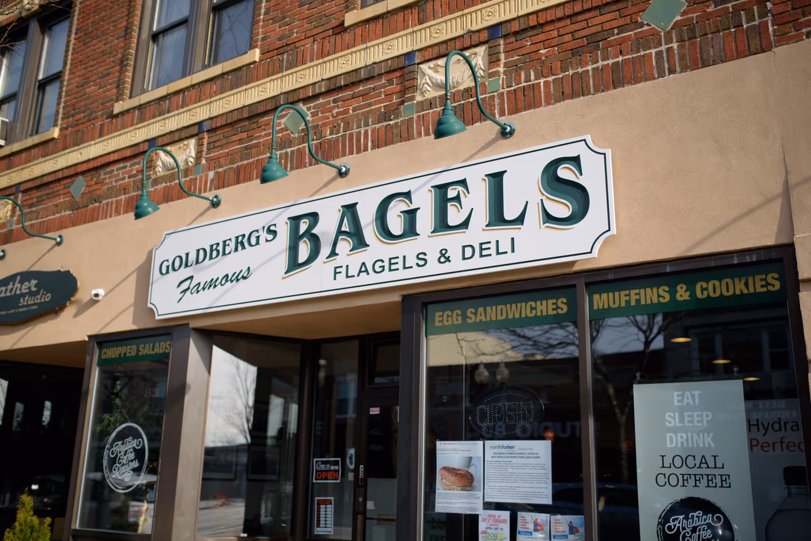 Exterior storefront of Goldberg's Bagels with a large hanging sign above windows advertising egg sandwiches, muffins, and local coffee.