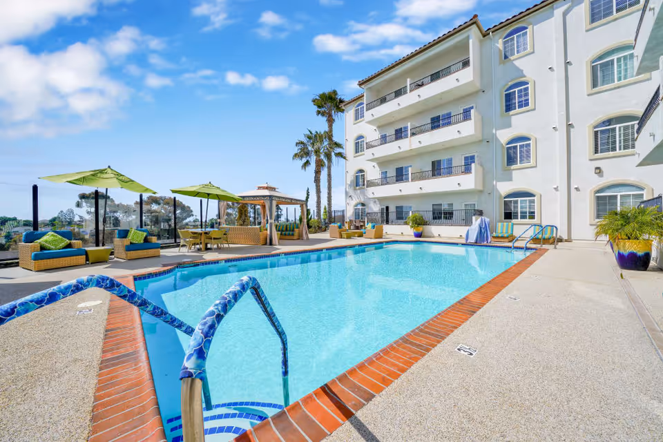 Outdoor swimming pool with lounge seating, green umbrellas, and a multi-story white building with balconies under a blue sky.