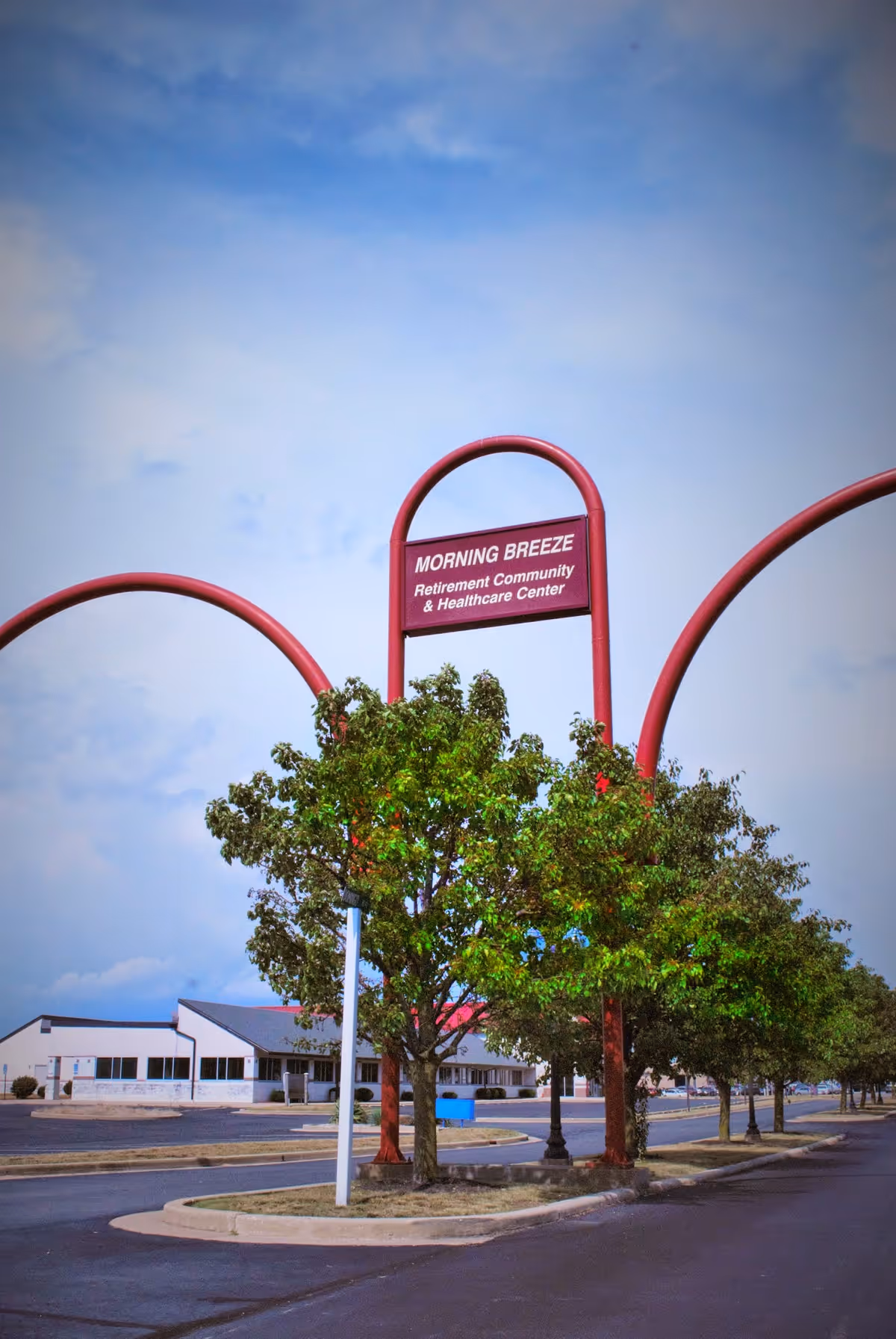Outdoor view of the Morning Breeze Retirement Community & Healthcare Center sign with red arches and trees lining the road, with the facility building visible in the background under a partly cloudy sky.