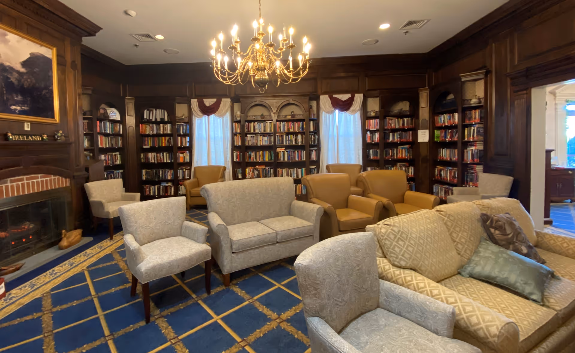 A cozy library room with wooden bookshelves filled with books lining the walls. The room features multiple armchairs and sofas arranged on a blue carpet with a gold pattern. A large chandelier hangs from the ceiling, and there is a fireplace on the left side with a painting above it. Two windows with curtains are visible in the background.