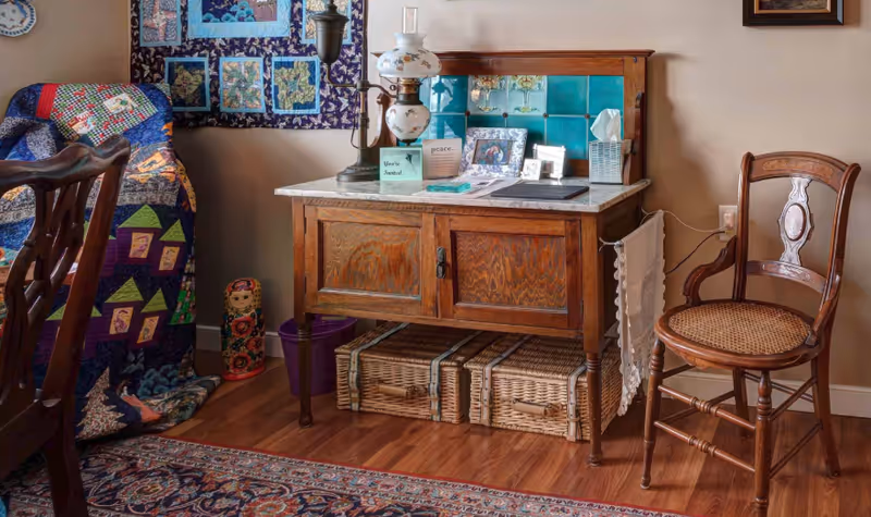 Cozy interior corner with a wooden sideboard topped with a lamp and framed photos, a woven chair, colorful quilt, and wicker baskets on a hardwood floor.