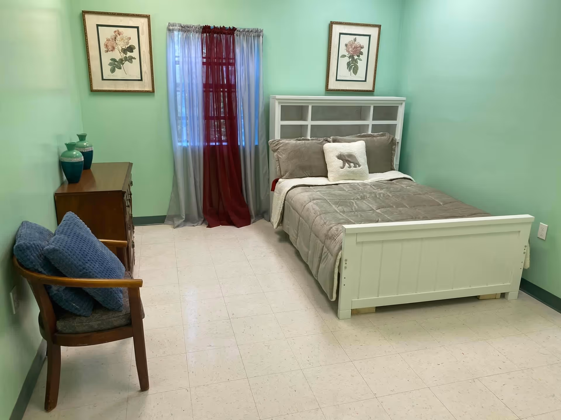 A small bedroom with light green walls and a white bed with gray bedding and a pillow featuring a bear. There is a wooden chair with two blue cushions on the left side and a wooden dresser with three green vases on top. The window has sheer gray curtains with a burgundy sheer curtain in the center. Two framed floral pictures hang on the walls above the dresser and the bed.