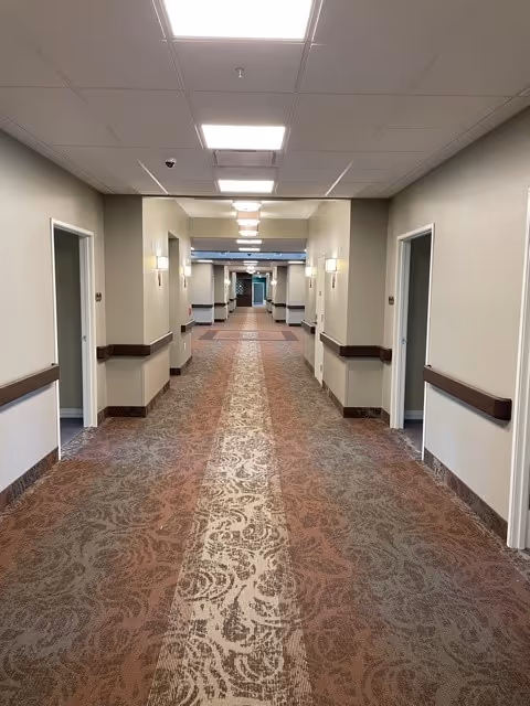 A long, well-lit hallway in a care and rehabilitation center with patterned carpet, beige walls, and multiple doorways on both sides. The ceiling has recessed lighting and there are handrails along the walls.